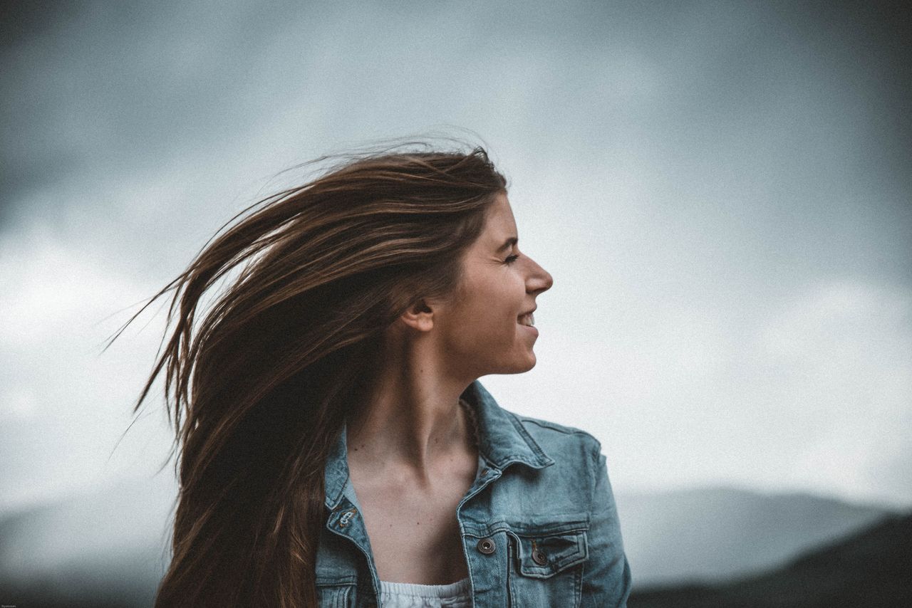 Portrait of a smiling young woman in a denim jacket against a cloudy sky.